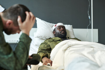 Young wounded African American soldier in military uniform lying in hospital bed while his worried friend sitting near by