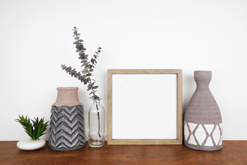 Mock up wood frame with clay vases, succulent plant and eucalyptus branch on a shelf or table. Wood shelf and white wall. Portrait frame orientation.