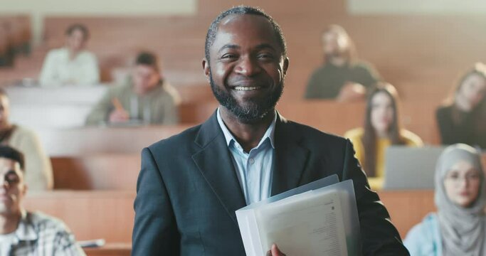Portrait of happy African American male professor smiling in front of camera and holding folder with papers. Students on background. Man teacher of high school. Camera zooming out. Close up.