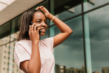 African american woman hold hand phone surfing internet chatting friends colleague call order taxi or food standing talking phone speaking outside street smiling happy positive good mood
