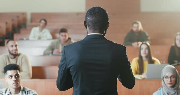 Rear of African American male professor standing in front of students in college and talking to them while having lection. High school. Back view. Man teacher turning to camera and smiling. Portrait.
