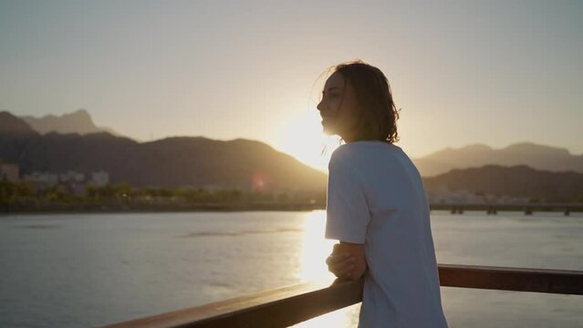 camera turning around happy carefree young woman standing at bridge railing, enjoying sunset time with orange sky. Sundown landscape with mountains and sea view