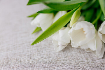 Obraz na płótnie Canvas An engagement ring in white gold with a diamond lies in a bouquet of white tulips on white background. Gift for Women's Day, Valentine's Day. Beautiful spring background with green leaves. Mock up