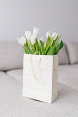 A bouquet of white tulips is in a gift bag on beige sofa in light interior. Background for Valentine's Day. Gift for Mother's Day or Women's Day. Spring background, March 8th	
