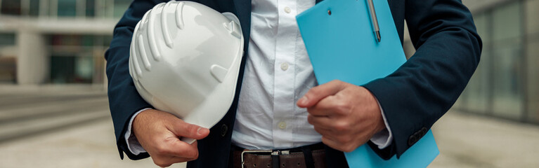 Close up of engineer in suit holding protective helmet and paper folder. Industrial concept