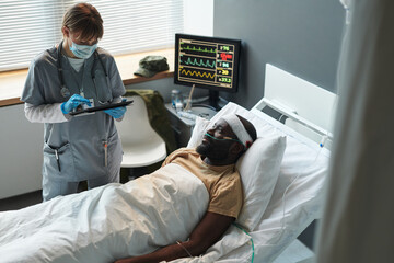 Obraz premium Young nurse in uniform and protective mask entering data in tablet while standing by patient with wounded head wrapped with bandage
