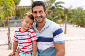 Portrait of happy biracial father and son embracing and smiling at beach - obrazy, fototapety, plakaty