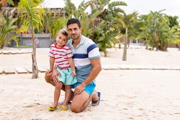 Portrait of happy biracial father and son embracing and smiling at beach, with slow motion - obrazy, fototapety, plakaty