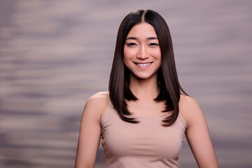 Fototapeta na wymiar Smiling asian woman wearing beige top and nude natural makeup portrait. Pretty young beauty model with joyful facial expression and healthy glowing skin looking at camera in studio
