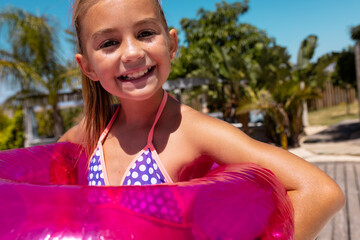 Portrait of happy biracial girl smiling and holding swim ring by the swimming pool - obrazy, fototapety, plakaty