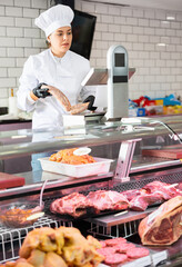 Positive young butcher shop saleswoman in white uniform working at counter in butchery, weighing fresh raw sausages on scale
