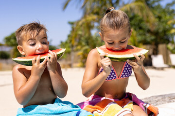 Happy biracial brother and sister eating watermelon by the swimming pool