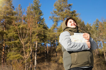 Photo from below of a traveler woman in winter clothes with a hood on her head looking at the sun and holding a map for trip in her hands.