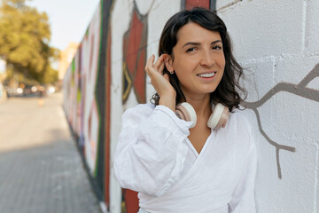 Charming cute lady with happy smile and short dark hairstyle wearing white shirt posing to camera, touching her hair and resting outdoors in sunny day over white wall in the city.