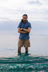 Young latin man looking at the camera with crossed arms and a bad face wading in the ocean. Vertical photo.