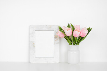 Mock up marble frame with pink spring tulip flowers. White shelf against a white wall. Copy space.