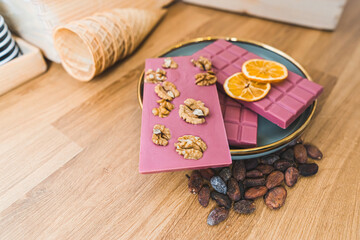 Various ruby pink chocolate bars with nuts, dried orange slices and toasted cacao beans on plate. Wooden table. High quality photo