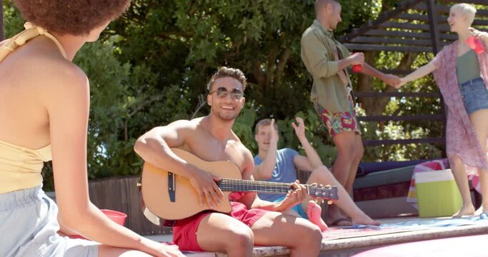 Happy diverse group of friends with drinks playing guitar at pool party in summer