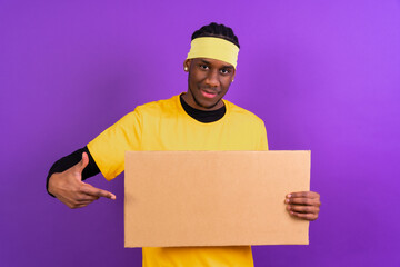 Black ethnic man in yellow clothes on a purple background, pointing holding a sign