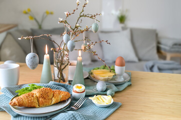 Breakfast table with boiled egg, croissant and pastel turquoise Easter decoration in a small living room, copy space, selected focus