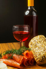 Still life with rolled smoked salmon, red wine, baby tomato, sliced onion and bread roll with seeds on a wooden board, dark background.