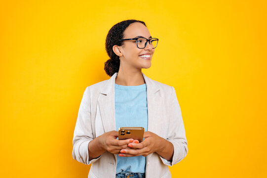 Lovely confident mixed race woman with glasses, stylishly dressed, using mobile phone, chatting in social media, texting message, looks to the side, smiling, stand on isolated yellow background