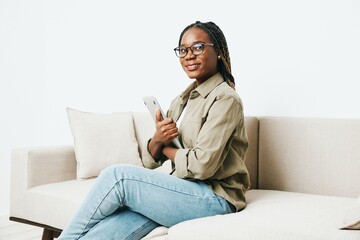 Obraz premium Stylish African American woman business freelancer working sitting on couch at home in a laptop and phone, business calls and correspondence, home clothes and eyeglasses, light interior background.