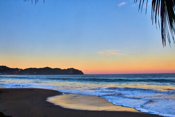 Fototapeta premium beach at sunrise with colors in the sky, coast in the background and palm tree in foreground, sayulita nayarit 