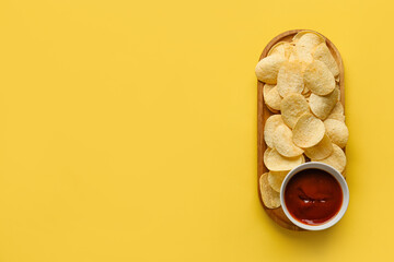 Board with delicious potato chips and bowl of ketchup on yellow background