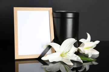 Blank funeral frame, urn and beautiful lily flowers on dark background