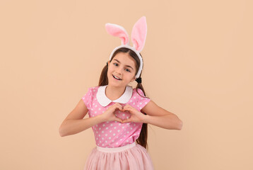 Obraz na płótnie Canvas Little girl in bunny ears making heart shape with hands on beige background