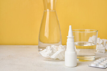 Nasal drops with pills, glass of water and tissues on table near yellow wall. Seasonal allergy concept
