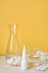 Nasal drops with pills, glass of water and tissues on table near yellow wall. Seasonal allergy concept
