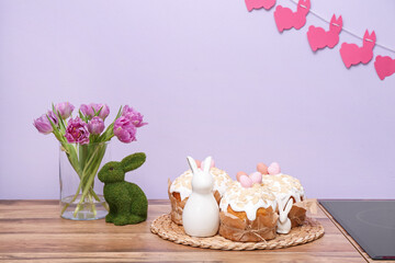 Easter cakes, bunnies and vase with tulip flowers on kitchen counter near lilac wall