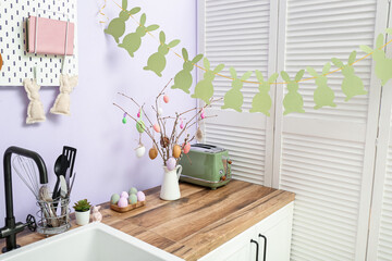 Vase with willow branches, painted eggs and bunny garland in kitchen decorated for Easter celebration