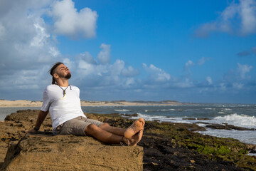 Young man sitting on a rock on a beach with an expression of happiness and relaxation.