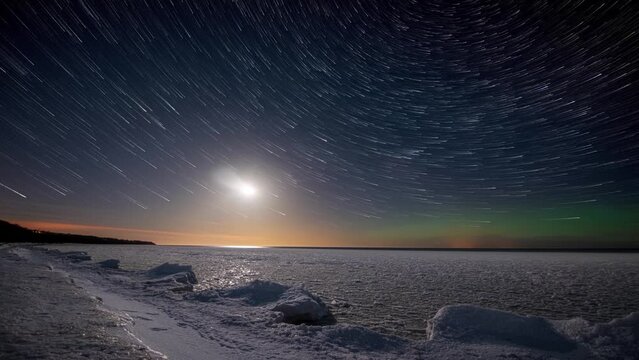 Timelapse. Star trails, moon trail and northern lights above the seashore and sea covered with ice.