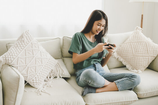 Happy young asian woman relaxing at home she is sitting on sofa and using mobile smartphone. She is playing online gaming have fun