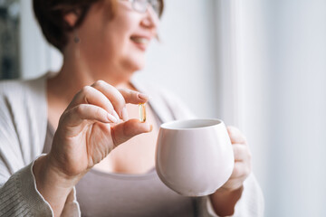 Close up photo of middle aged woman holding omega 3 capsule and mug of water in hands near window...