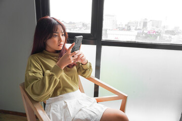 A young Asian woman is feeling serious and focused on holding her smartphone in hand while sitting in a bedroom.