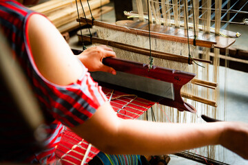 Horizontal photo from behind of the hands of a silk weaver working on her loom on a red piece of...