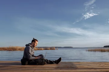 Foto op Canvas Young male working online on a laptop while sitting on a wooden pier. Freelancer using laptop for internet work outside. The concept of remote work. © Kaplitskaya Love