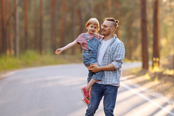 Fototapeta premium A magical moment unfolds as a father holds little daughter in his arms and they laugh joyously while strolling along a forest road, surrounded by majestic pine trees and the warm glow of the sunset.
