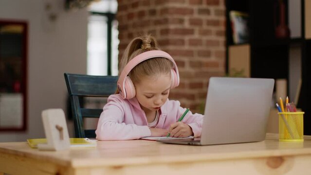 Adorable little schoolgirl in headset studying online at home, looking at task on laptop screen and writing in copy book
