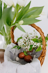 A wicker basket, on a gray linen napkin, are painted eggs and white branches with blossoming pear flowers, an Easter wooden rabbit an glass vase with white fresh tulips. Spring Easter background