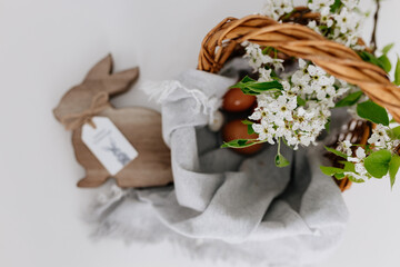 Fototapeta na wymiar A wicker basket, inside which, on a gray linen napkin, are painted eggs and white branches with blossoming pear flowers, next to it stands an Easter wooden rabbit. Spring background for Easter card