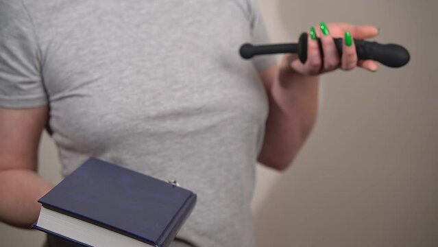 a woman in a gray t-shirt holds a black dildo and a book in her hands. choice between education and online freelancing. High quality Full HD video recording