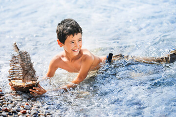 Happy child is playing with wooden toy ship in the sea water.