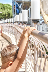 Blond boy with wet hair holds on to railing near big slide in water park on sunny day. Preschooler enjoys summer holiday at resort closeup