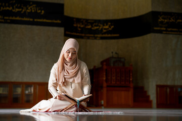 The image of an Asian Muslim woman in the Islamic religion in hijab in cream color. Sitting reading the Quran and having a happy smiling face Staying in a beautiful mosque out of respect for God.